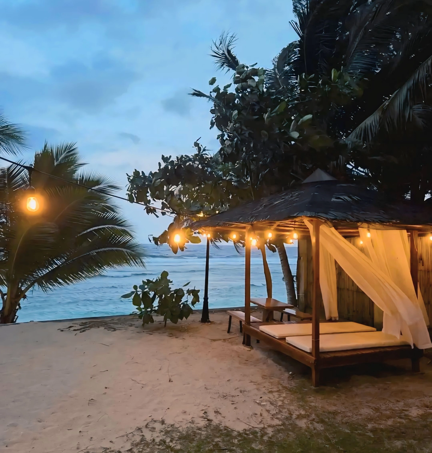 Beachfront cabana with fairy lights at dusk, palm trees, and ocean waves.